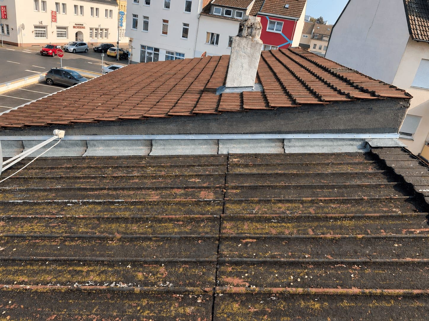 Verwittertes Dach mit bemoosten Wellplatten, dahinter ein altes Ziegeldach mit gemauertem Schornstein